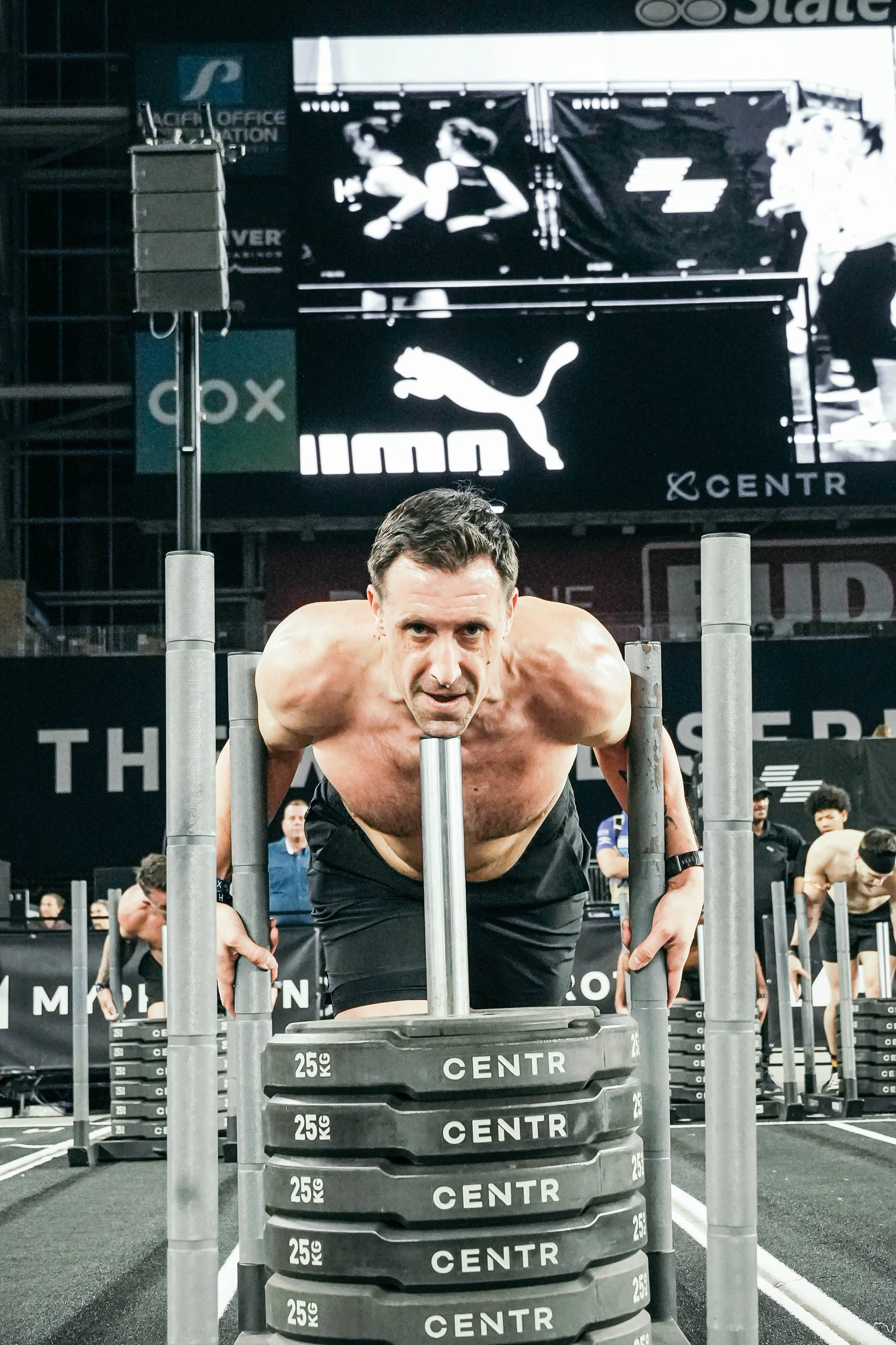 A shirtless man in black shorts doing a push-up on a stack of weight plates in a gym, with a large screen in the background displaying a wrestling match and Puma logos.