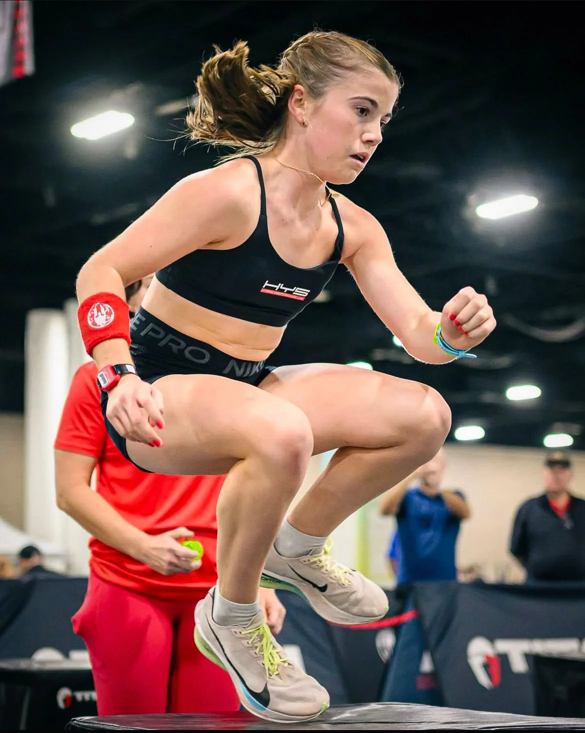 Female athlete jumping on a fitness platform during an indoor competition, with onlookers in the background.