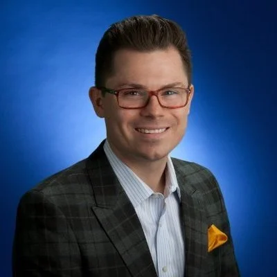 Man wearing glasses and a plaid suit jacket with a yellow pocket square, smiling against a blue background.