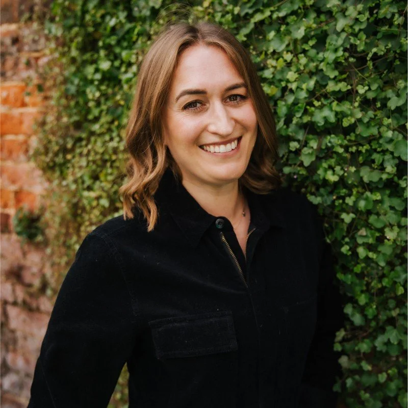 Woman smiling in front of green ivy-covered wall, wearing a black jacket, with a brick wall partially visible.