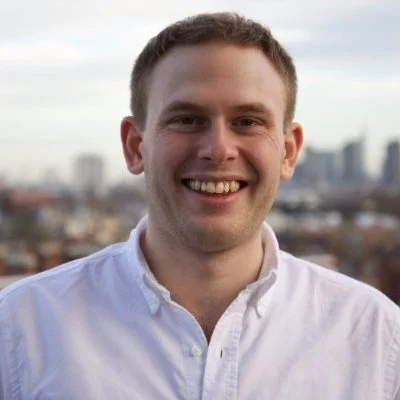 A smiling man in a white button-up shirt standing outdoors with a city skyline in the background.