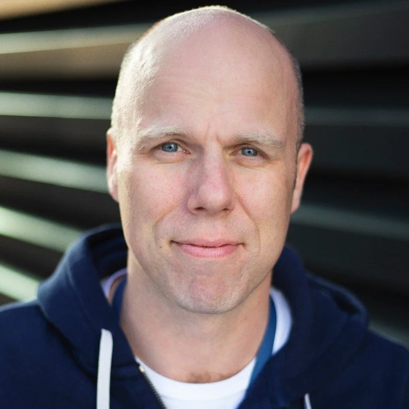 Close-up of a middle-aged man with a shaved head and blue eyes, wearing a navy hoodie and standing outdoors with a blurred background of horizontal metal or plastic slats.