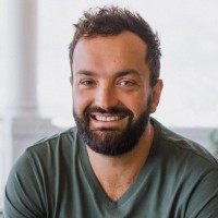 Smiling man with a beard and short curly hair, wearing a green shirt, indoors with a windowed cityscape background.
