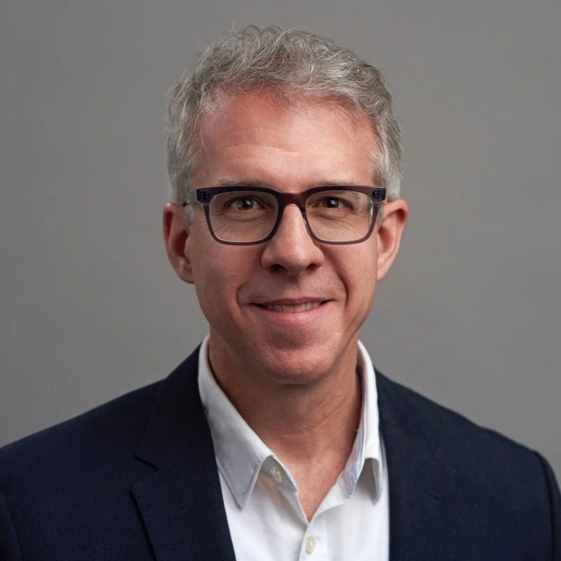 Portrait of a smiling man wearing glasses, a dark blazer, and a white shirt, against a plain background.