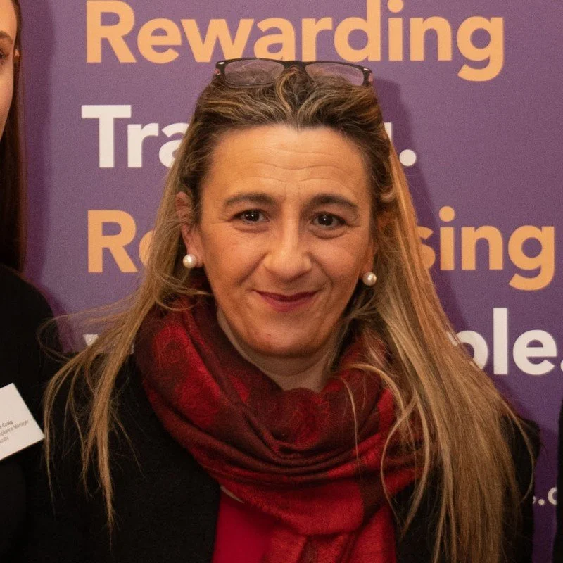 Woman with glasses on her head, wearing a red scarf and pearl earrings, standing in front of a purple background with partially visible text.