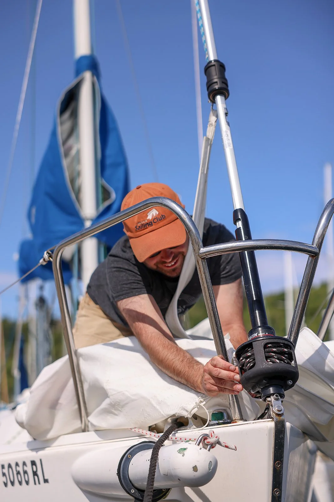 A man wearing an orange cap labeled 'Sailing Club' adjusts the front rigging of a sailboat with a blue sail in a clear outdoor setting.