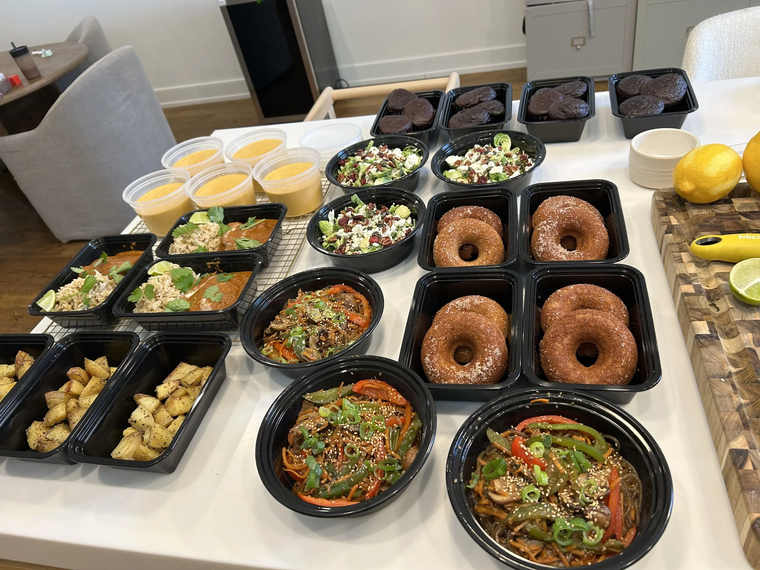 An assortment of food on a white table, including black containers with donuts, salads, rice dishes, roasted potatoes, and soups, with lemons, limes, and a yellow measuring tape on the side.