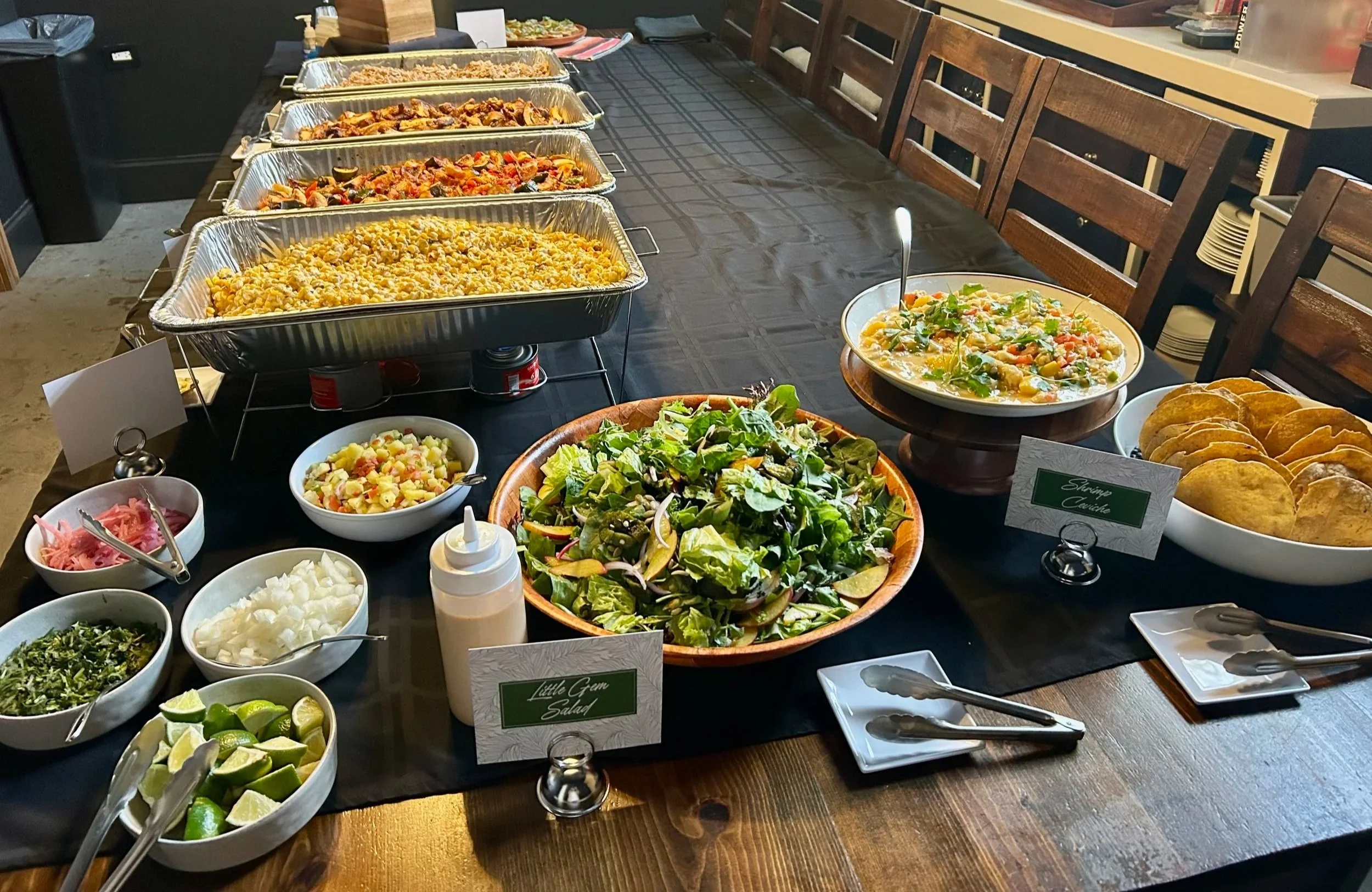 A buffet table with various foods including salads, fried snacks, pasta, and chicken dishes, with serving utensils and bowls of lime wedges and other condiments on a dark tablecloth in a dining area.