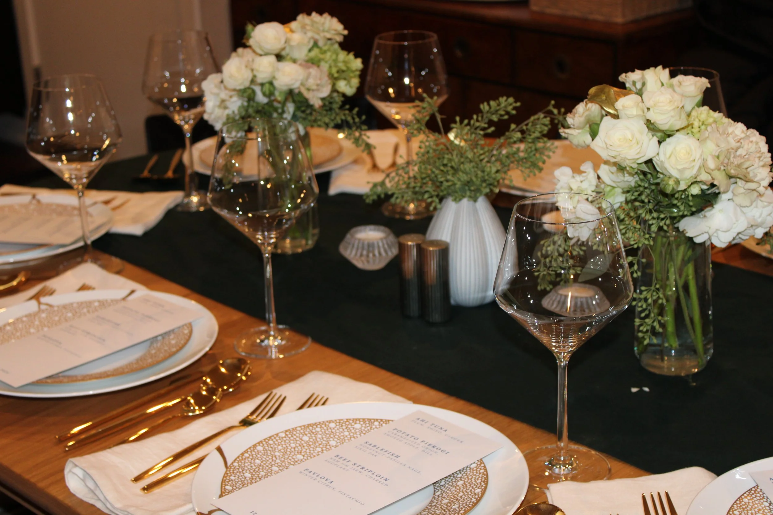 Elegant dining table decorated with white floral arrangements, wine glasses, and gold flatware on white napkins, set for a formal meal.