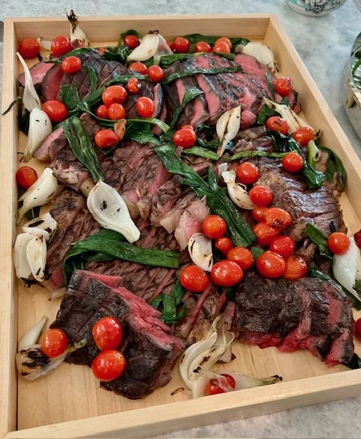 Cooked sliced beef, cherry tomatoes, roasted garlic, and green herbs on a wooden serving tray