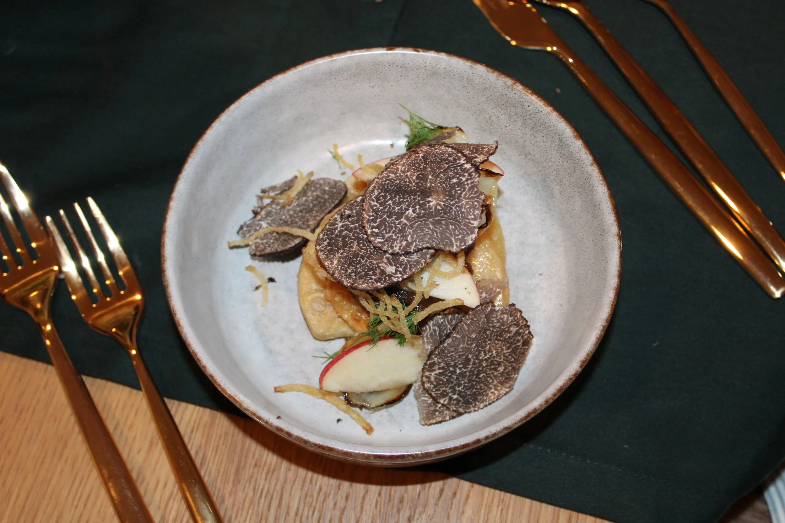 Dish with truffle slices, potatoes, and greens served on a ceramic plate, surrounded by gold utensils on a dark green and wood table.