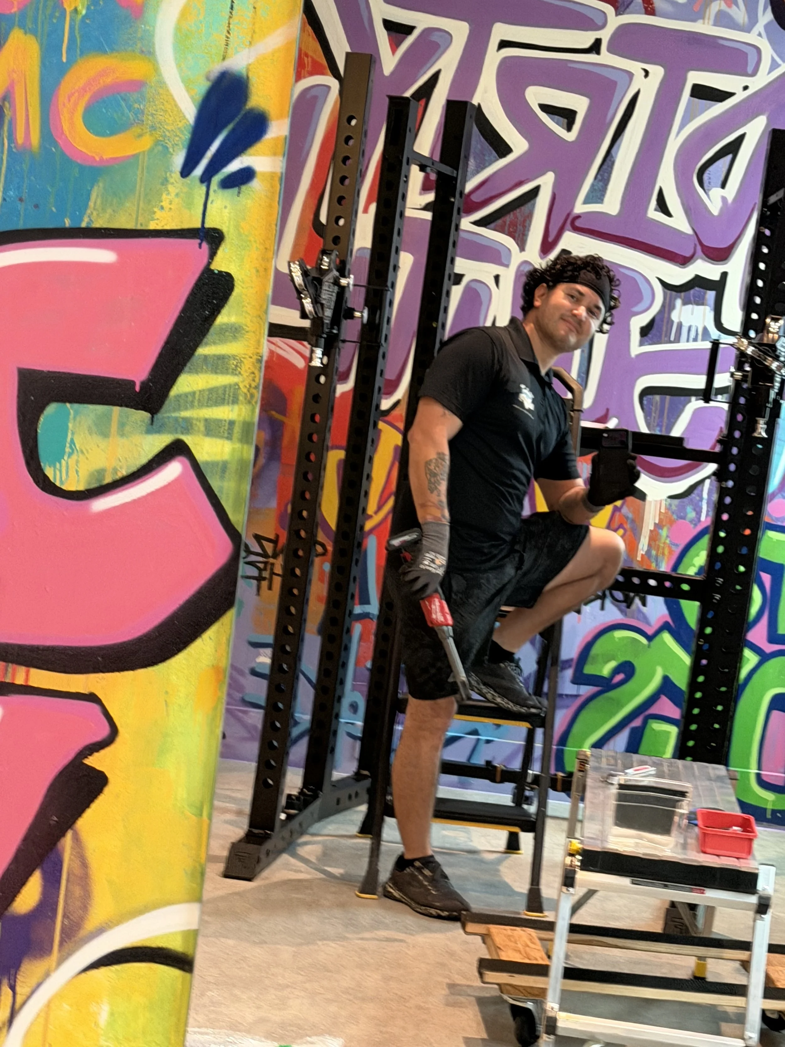 A man in black athletic clothing and gloves kneeling on one leg on a chair, holding a power drill in one hand, inside a room with colorful graffiti on the walls.