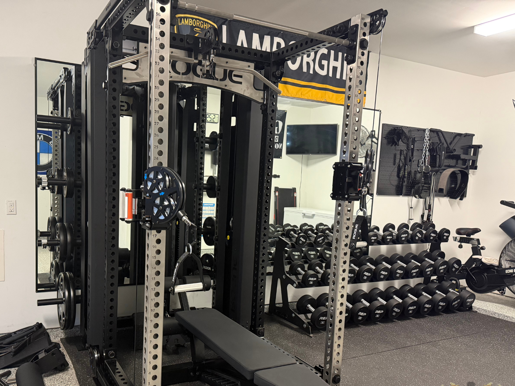 A gym with black weightlifting equipment, a rack of dumbbells, a mirror, and a TV on the wall.