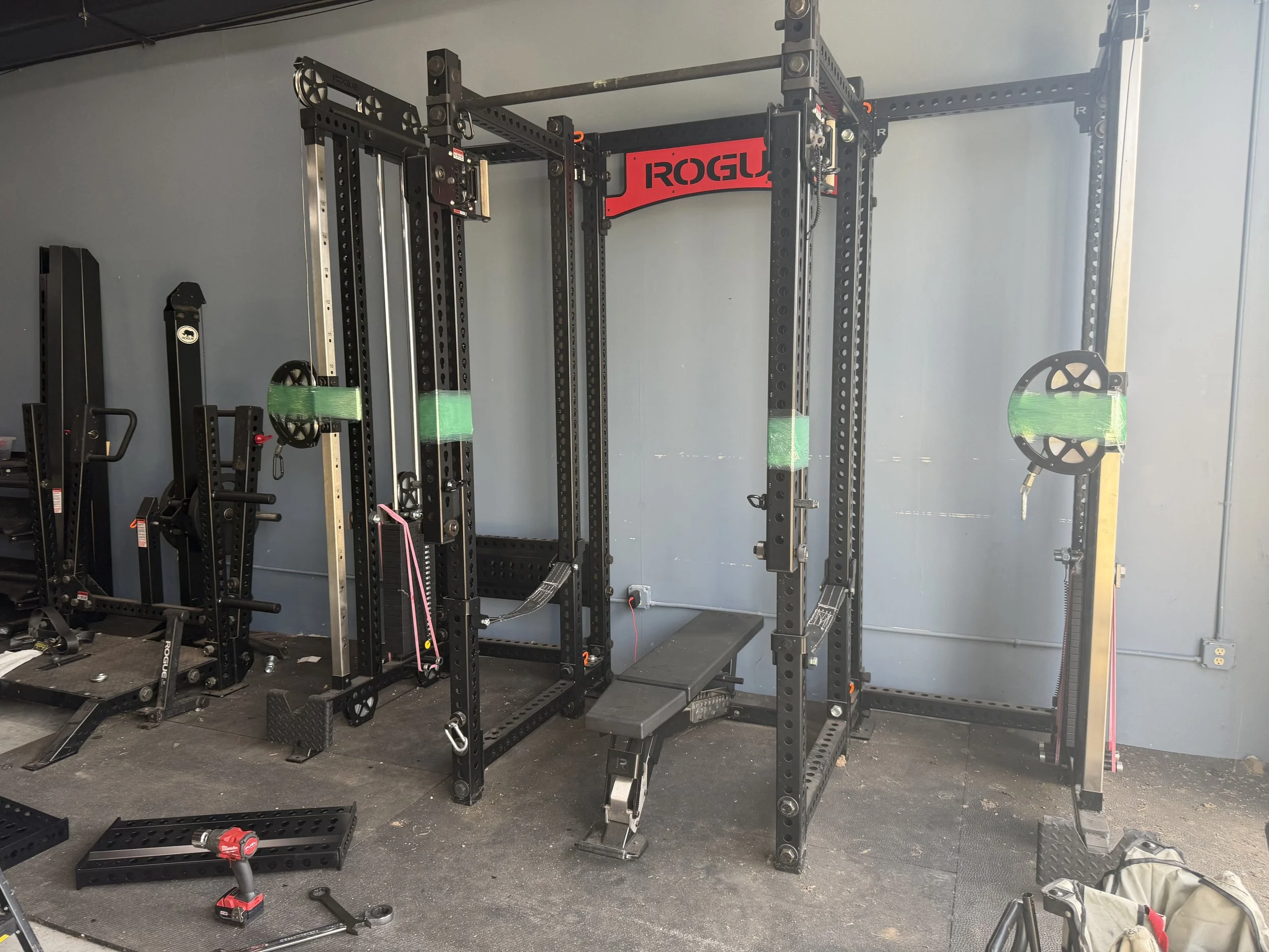 A partially assembled black metal power rack with red accents, including pull-up bars, weight storage, and accessories, in a gym. There are tools and parts on the floor nearby, and a gray wall with an electrical outlet behind the equipment.