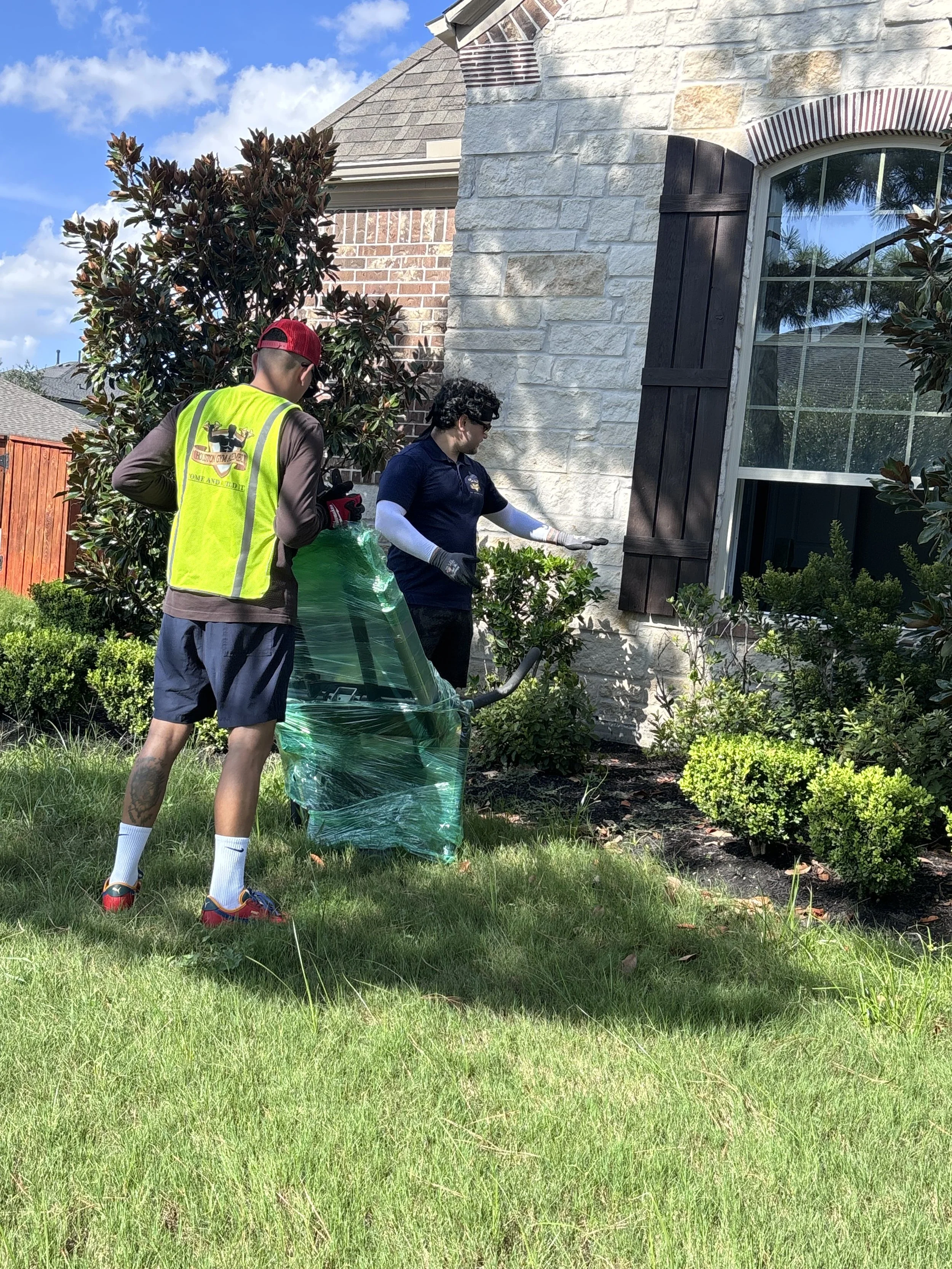 Two people are working in a garden next to a house with brick and stone exterior, a window with shutters, and bushes. One person is wearing a yellow safety vest, shorts, and a red cap, and the other is wearing a navy shirt and gloves, holding a garden tool.