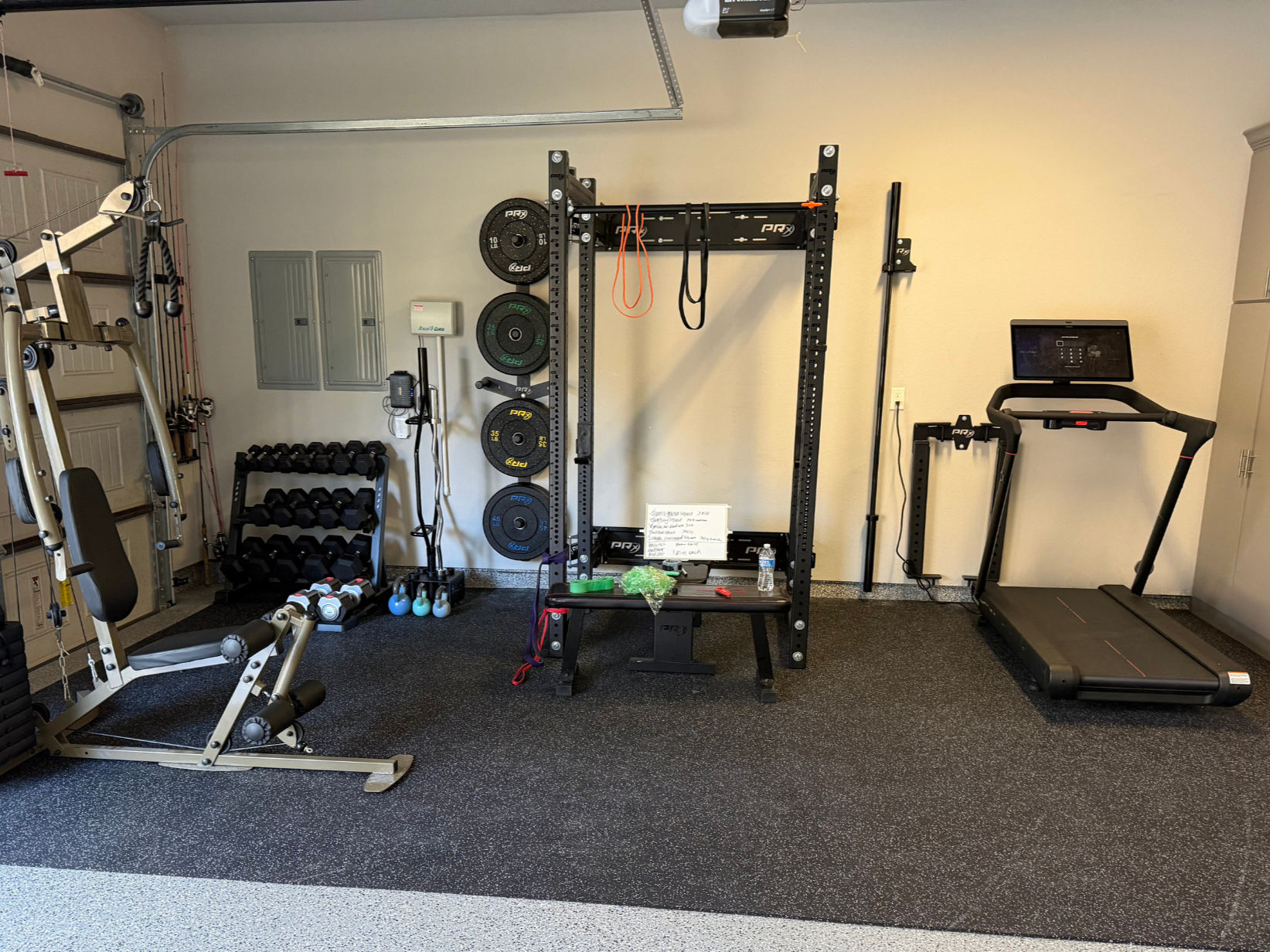 Home gym setup with various exercise equipment, including a treadmill, weight rack with weights, a power rack, dumbbells, kettlebells, resistance bands, and a whiteboard.