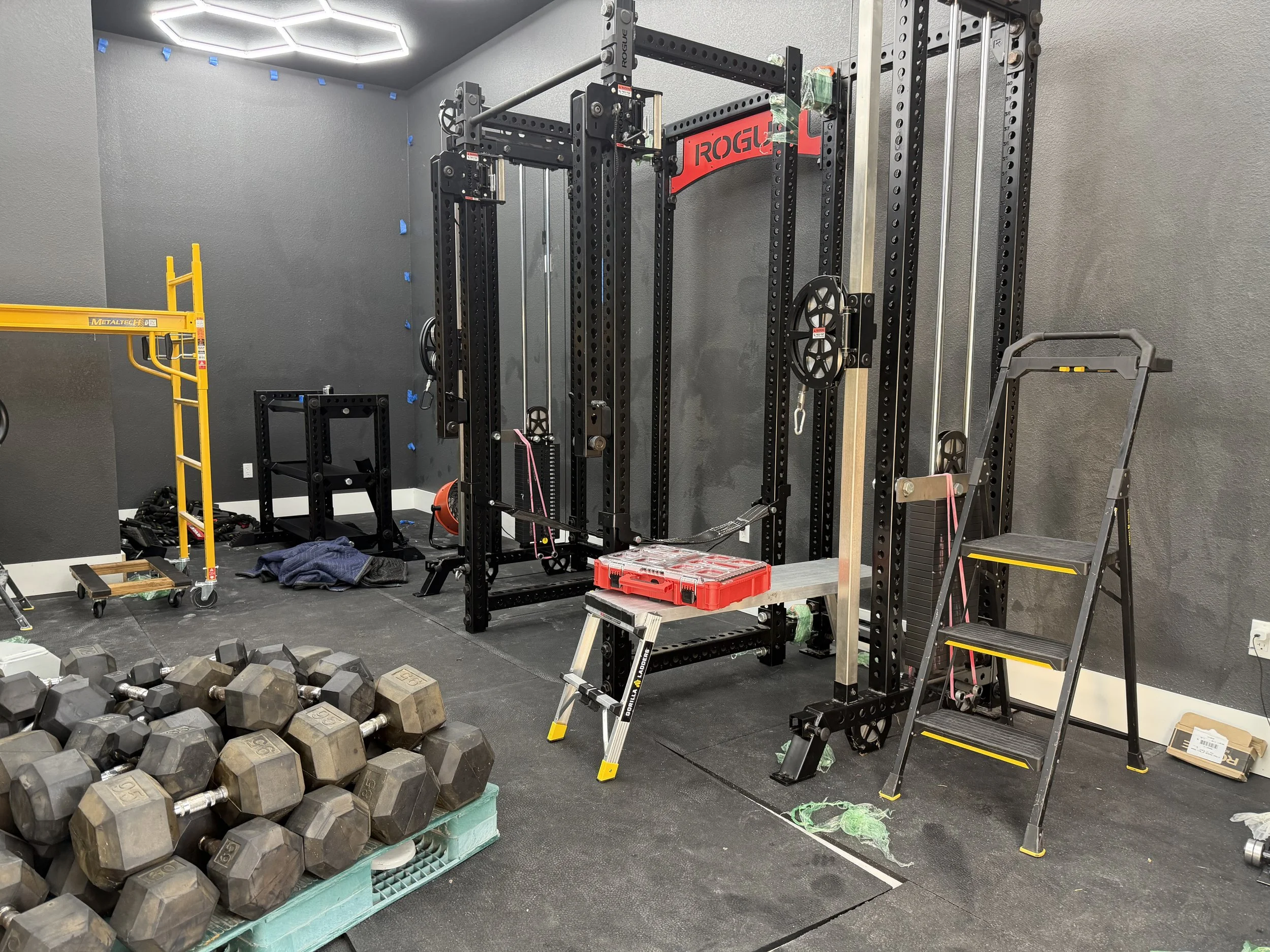 A gym room with various equipment, including a rack with weights, a ladder, a step platform, and scattered workout items.