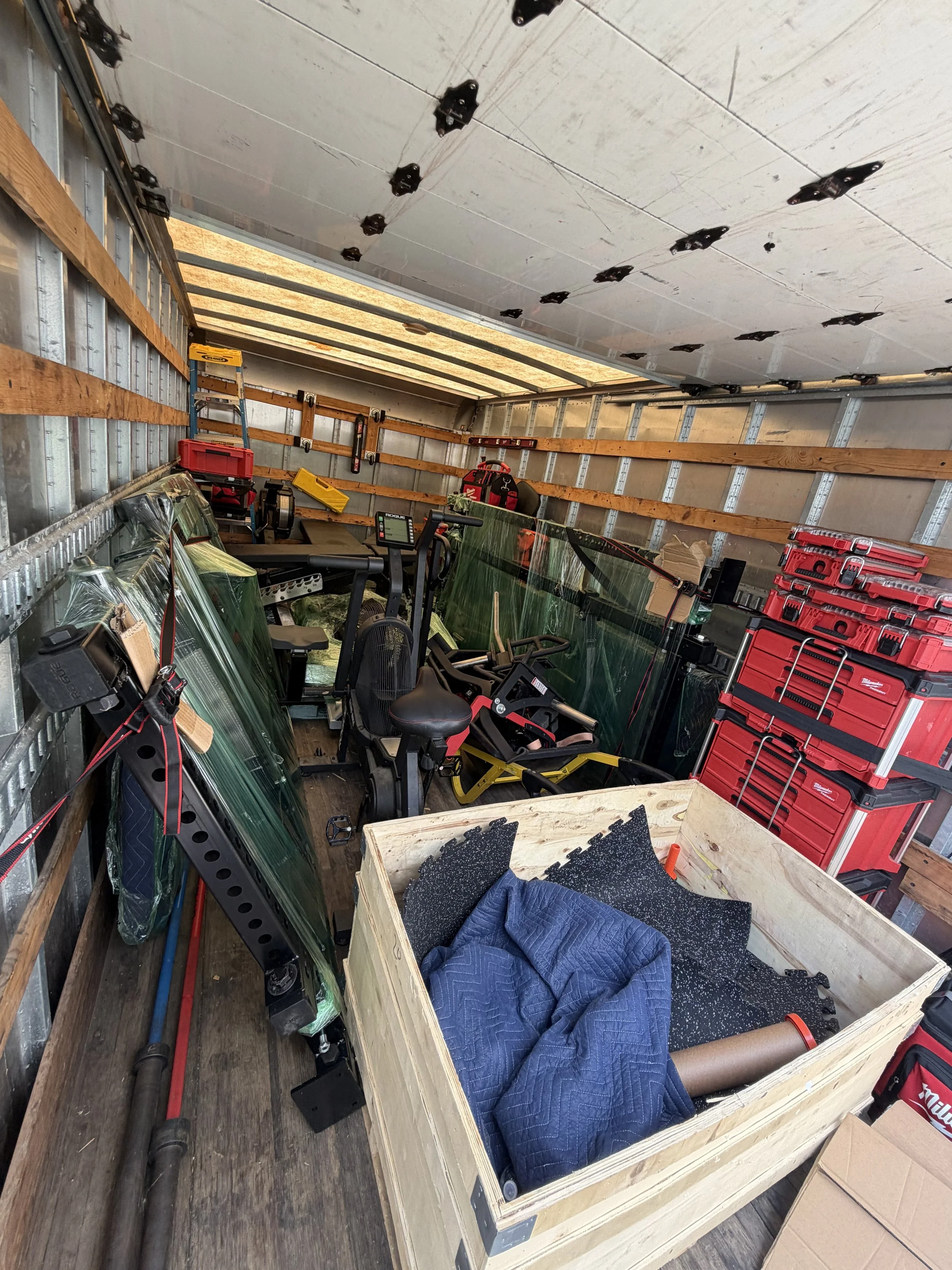 Interior of a moving truck with construction tools, plywood, stacked shingles, and a wooden box with a quilt and foam mat inside.