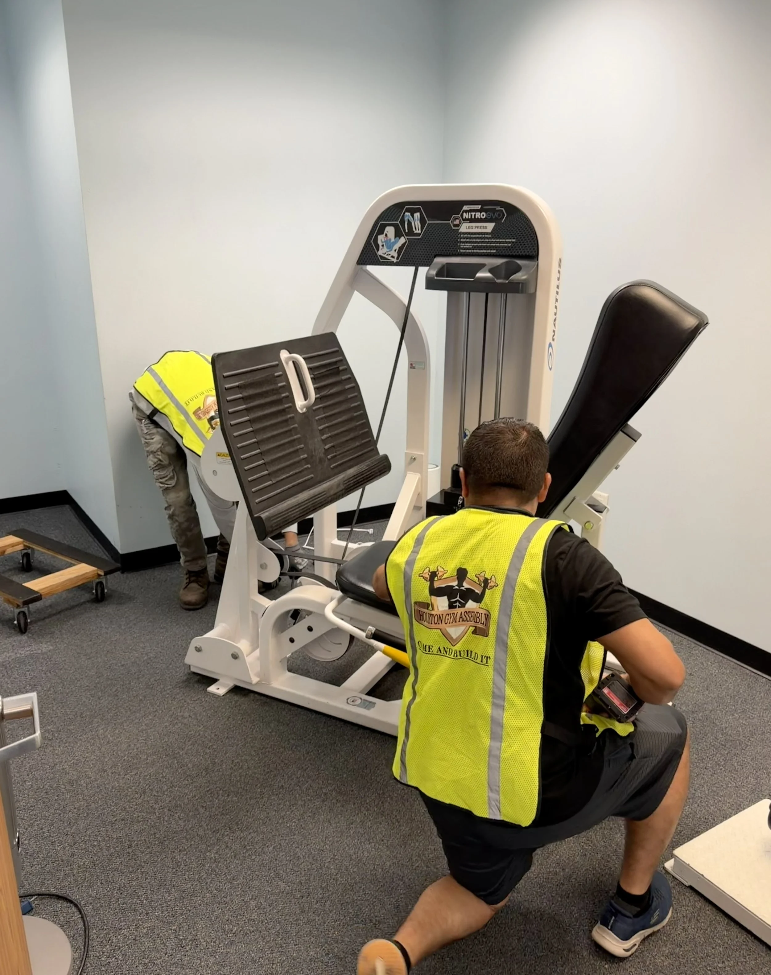 A person in a yellow reflective vest working on assembling gym equipment in a room with light blue walls and dark carpet flooring.