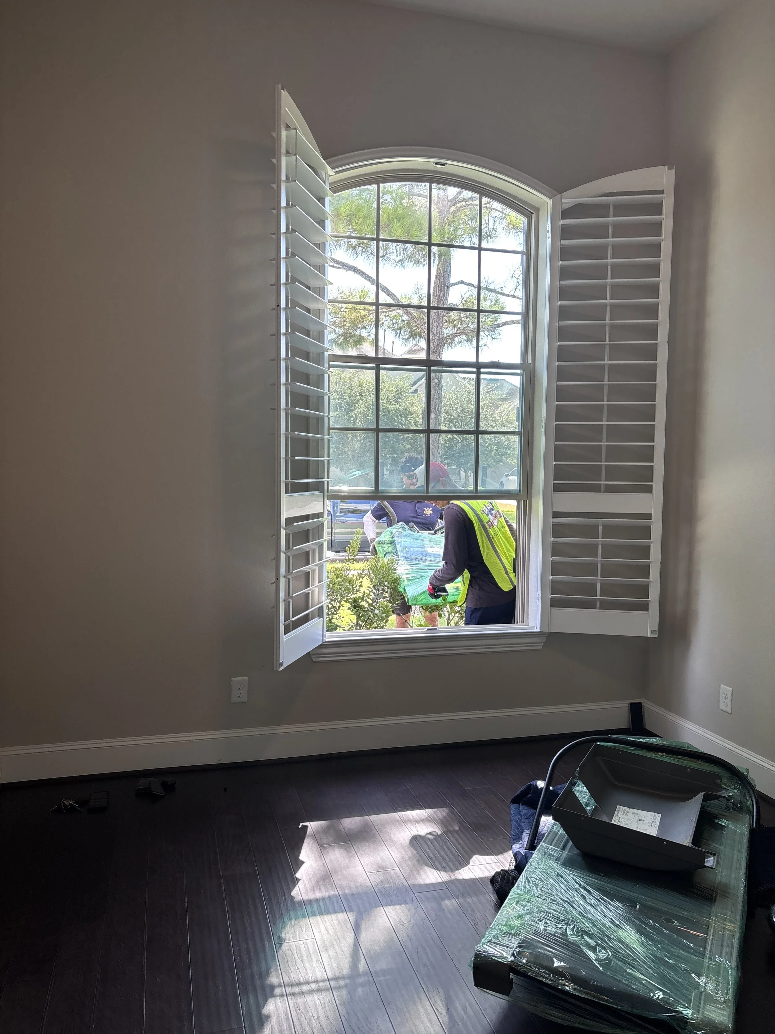 View from inside a room through open window shutters showing workers outside planting or working in a garden.