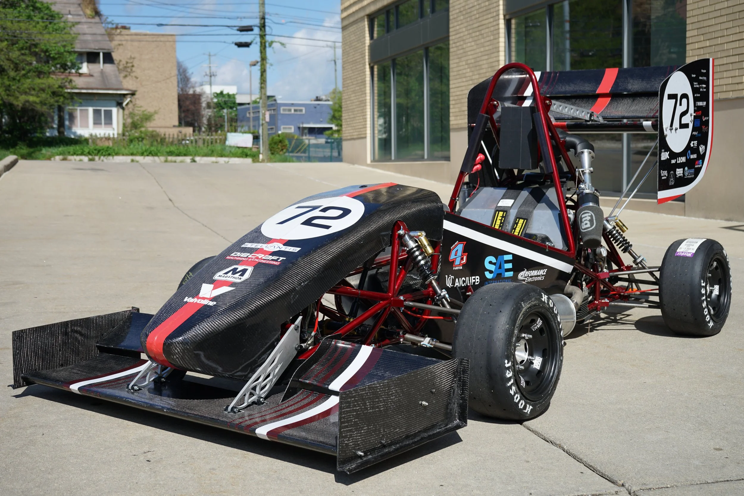A race car with a black and red design, number 72, parked on a concrete driveway with houses and trees in the background.
