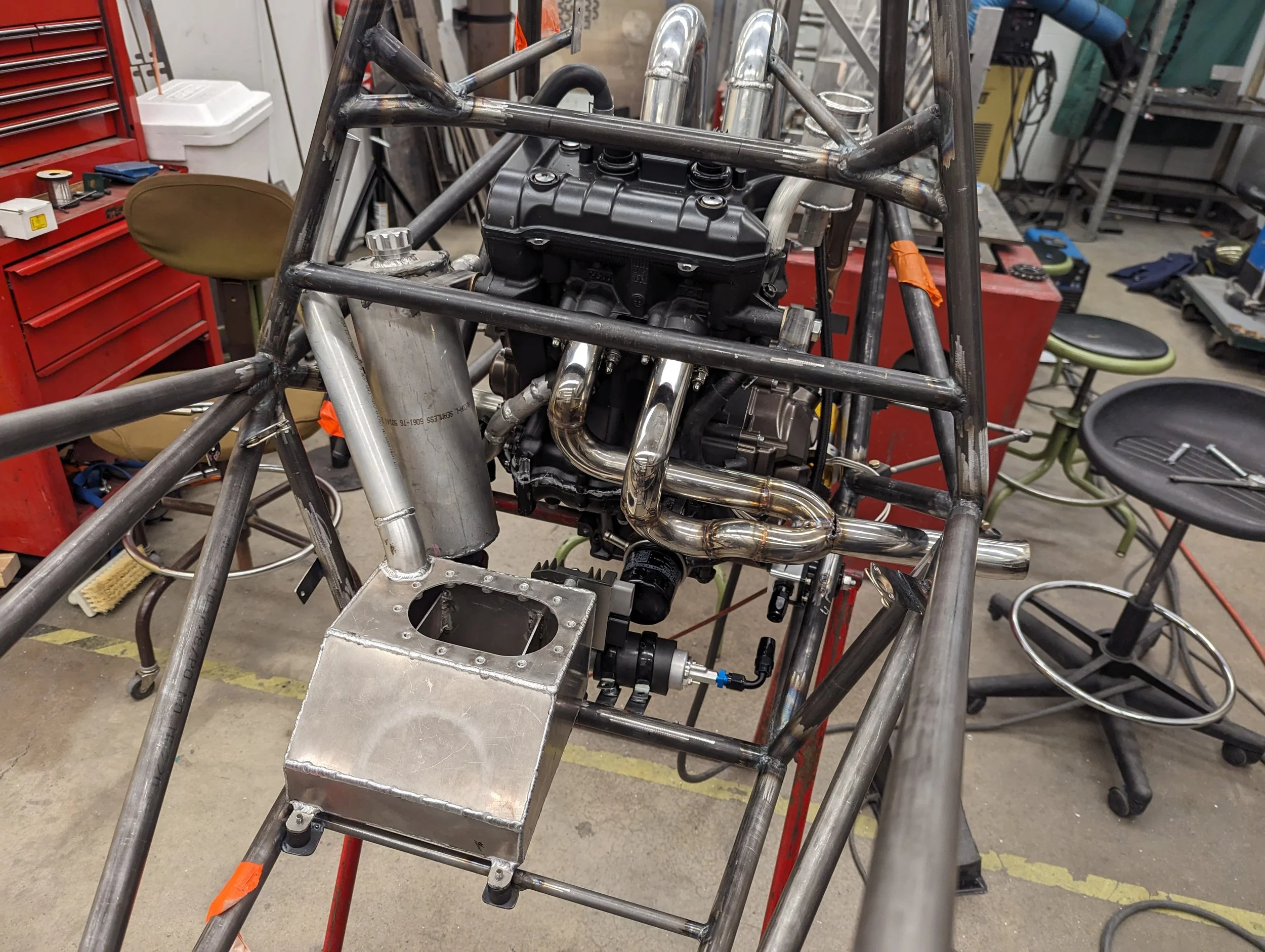 Interior of a workshop with a metal frame and a partially assembled racing car, with an engine, exhaust system, and aluminum components visible, surrounded by tools and stools.