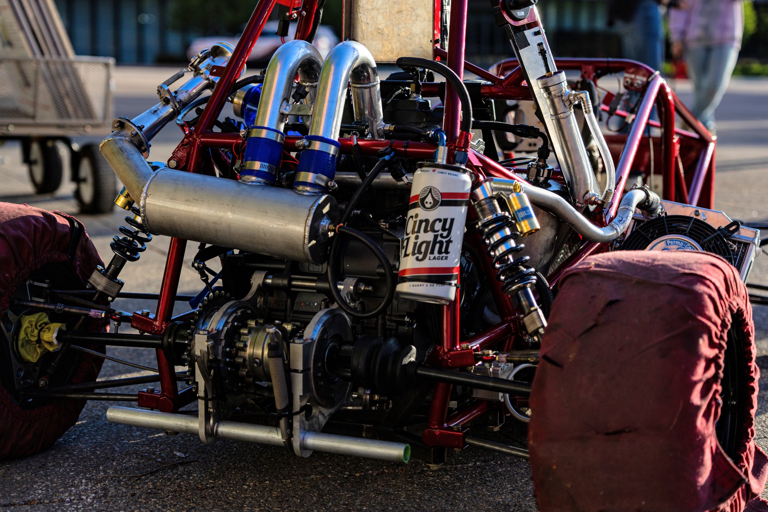 Close-up of a race car engine and chassis with red frame, metallic parts, and a Cincy Light Lager beer can attached. The tires are covered with protective cloths. Background shows a person and a cart.