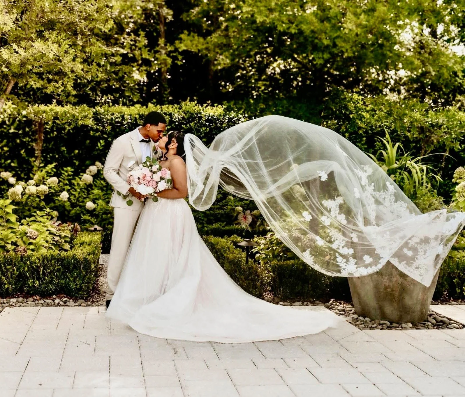 A bride and groom in wedding attire sharing an intimate moment outdoors, with the bride holding a bouquet of pink and white flowers, and a flowing veil in the air. Greenery and flowering bushes surround them.