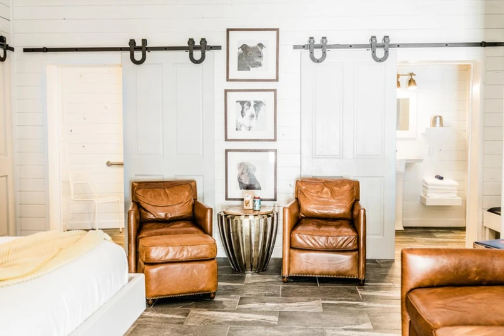 Modern rustic room with leather armchairs, framed animal art, barn-style sliding doors, and visible bathroom. Features white walls and wood-style flooring.
