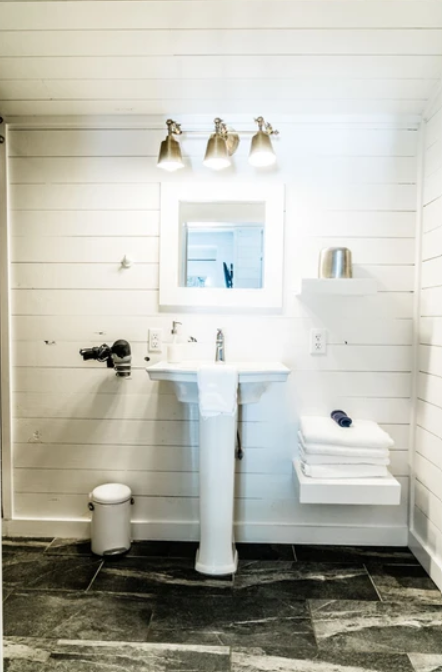 White bathroom with pedestal sink, mirror, and overhead lights. Towels stacked on a floating shelf, small trash can on tiled floor.
