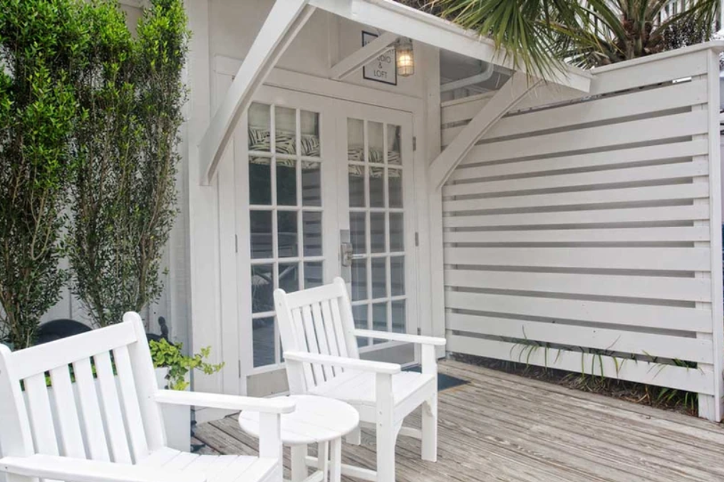 White patio featuring two white wooden chairs and a small table on a wooden deck, in front of a building with French doors and white siding. Tall plants provide partial privacy on one side.