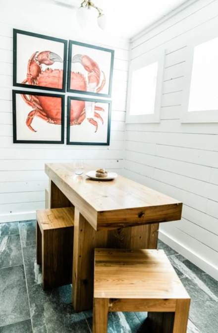 A minimalist dining area with a wooden table and benches on a tiled floor. The white walls feature a four-piece framed artwork of a red crab. Two windows provide natural light.