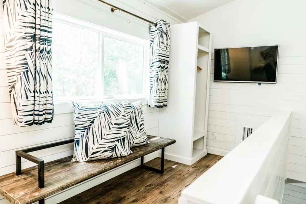 A modern room with a rustic wooden bench, two pillows with black and white leaf patterns, a wall-mounted TV, and a large window with matching curtains.