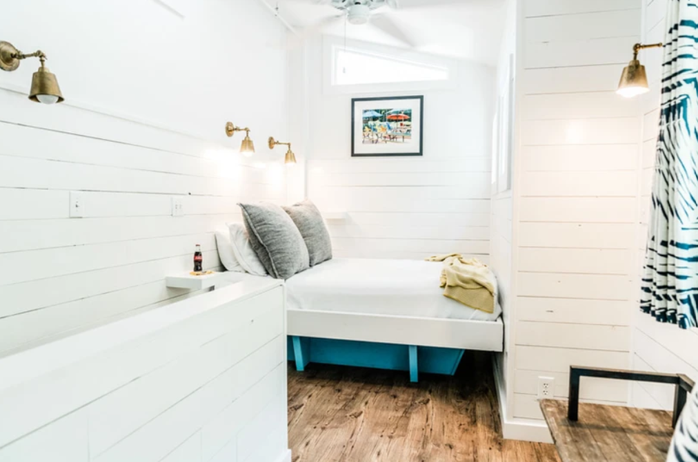 Small bedroom with white shiplap walls, a cozy bed with gray pillows and a yellow blanket, wooden floor, brass wall sconces, a framed picture, and a zebra-print curtain.