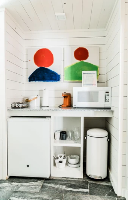 Small kitchenette with microwave, mini-fridge, and countertop appliances, featuring two abstract paintings of red circles, one with a blue shape and another with a green shape beneath. The space includes shelves with dishes and glasses, and a white t