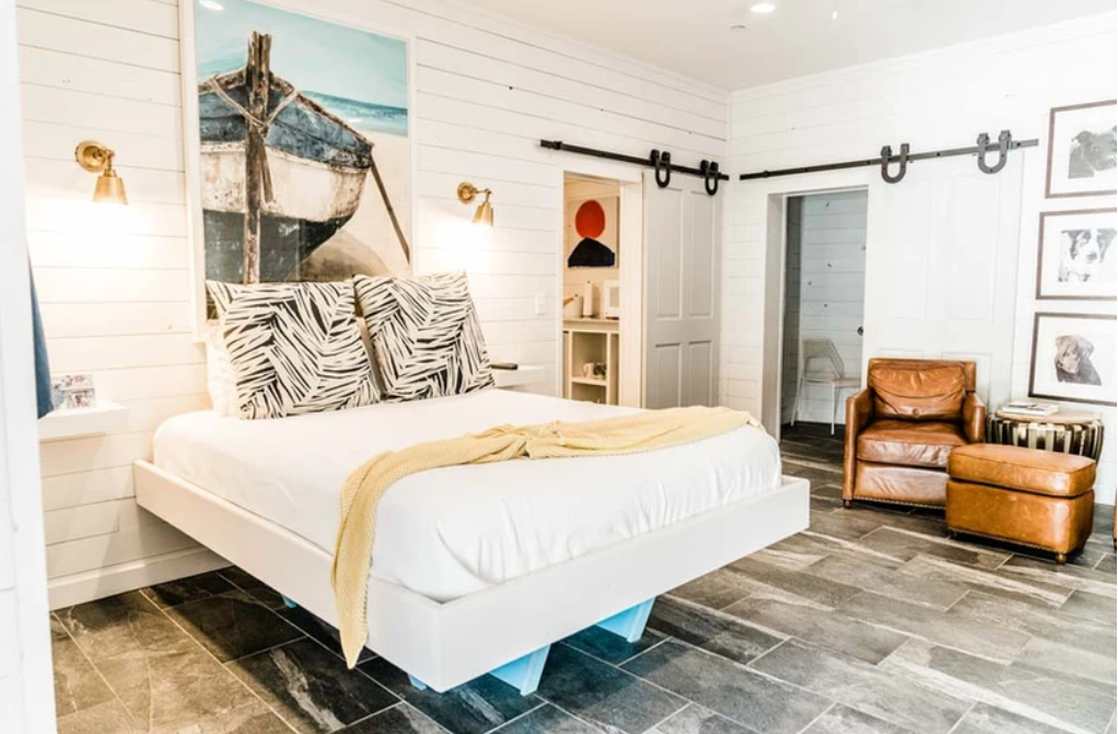 Modern bedroom with white shiplap walls, a floating bed with tropical-themed pillows, a yellow throw blanket, and nautical artwork above. The room features two sliding barn doors, brown leather armchair and ottoman, and black-and-white portraits on t