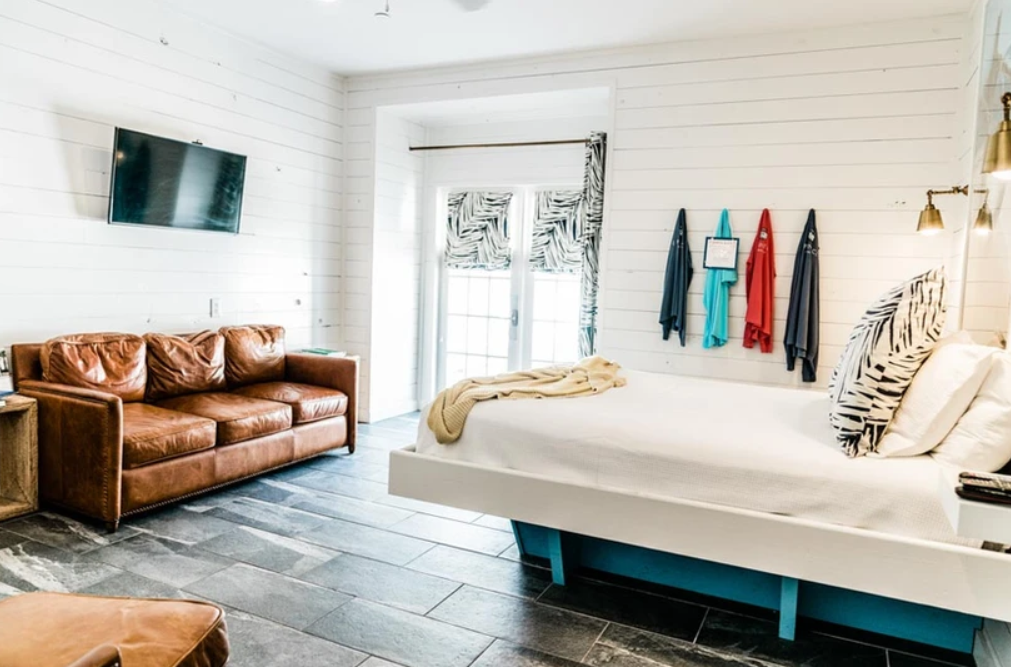 Stylish bedroom with leather sofa, wall-mounted TV, bed with decorative pillows, hanging clothing, wood panel walls, and zebra-patterned curtains.