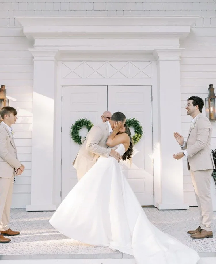 Bride and groom kissing at wedding ceremony with officiant and groomsmen on porch of a white building.