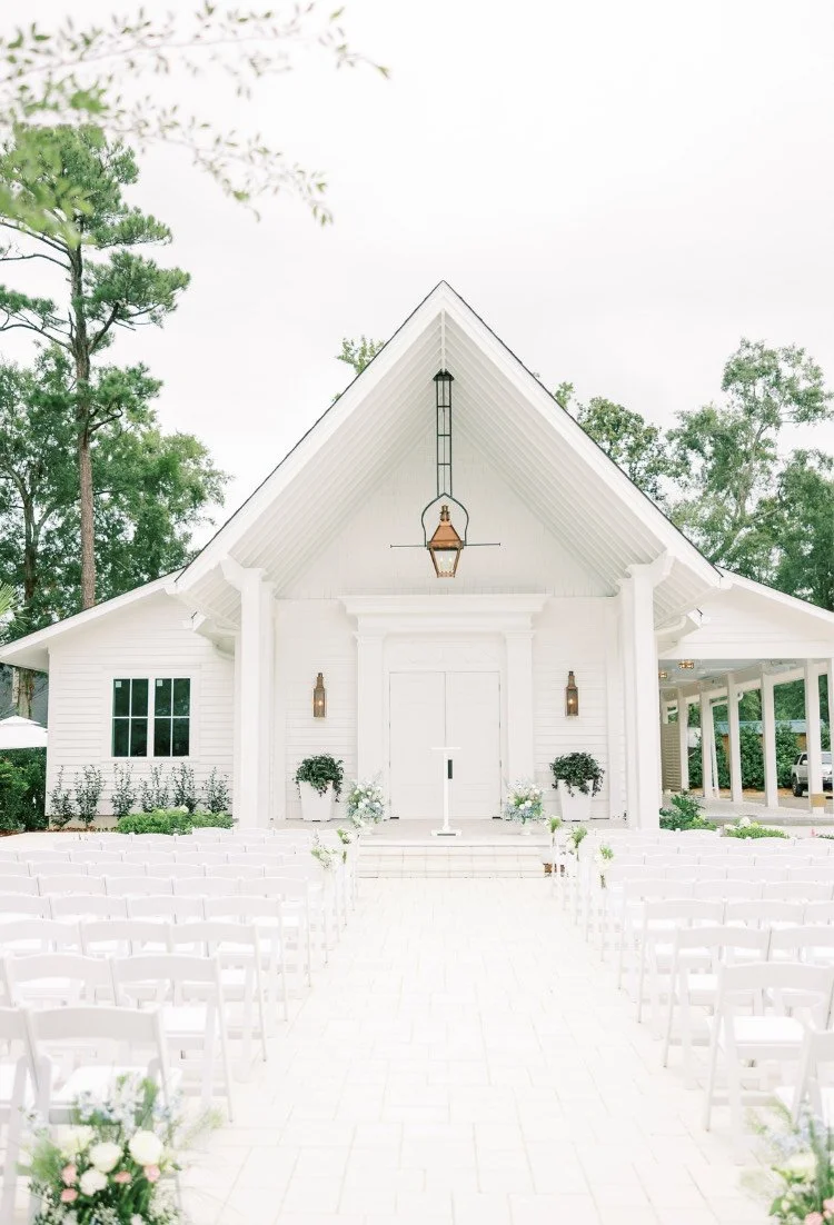 White chapel with outdoor seating arranged for a wedding ceremony, surrounded by trees.