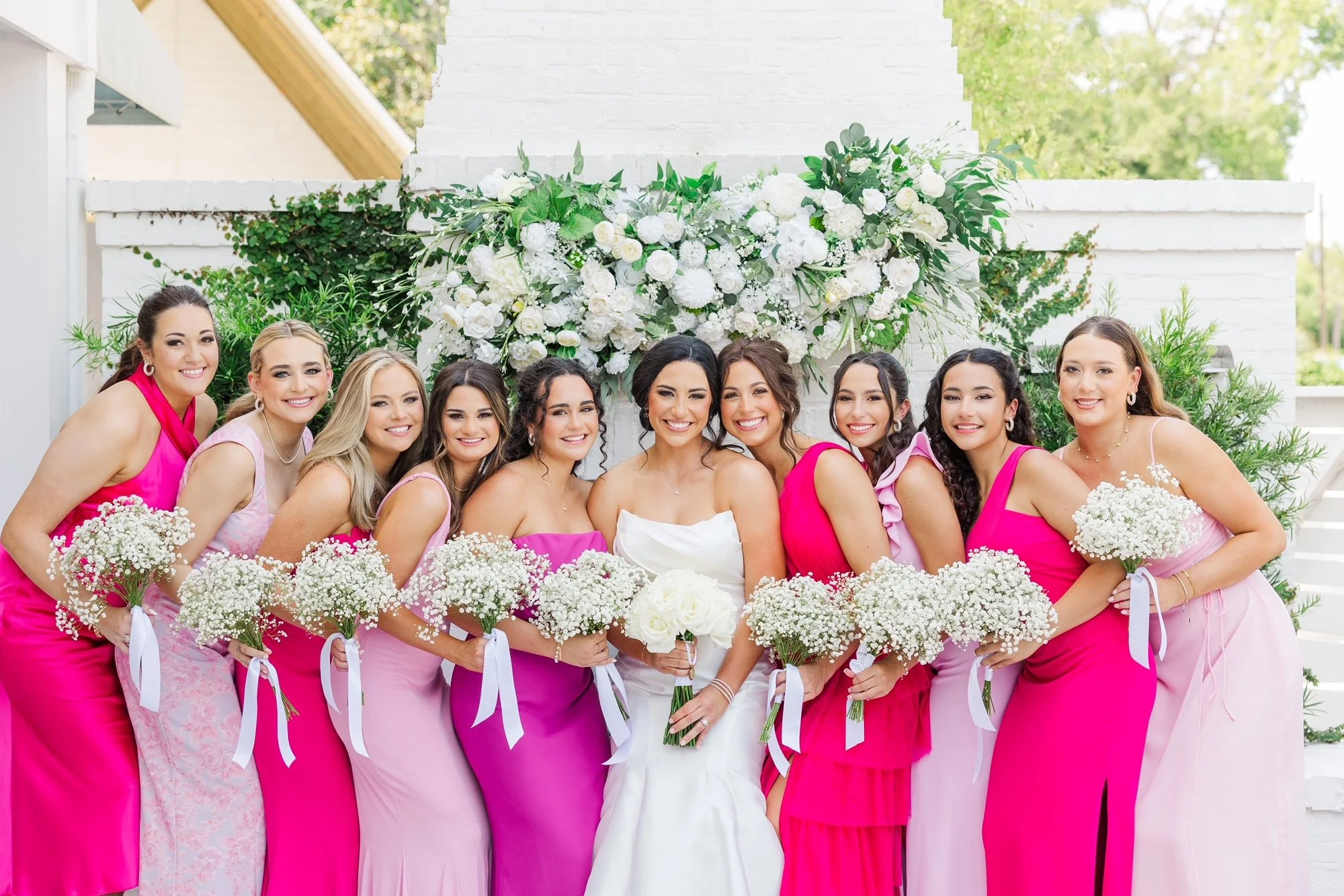 A bride in a white gown poses with her bridesmaids, who are wearing pink dresses, in front of a floral backdrop. The bridesmaids hold bouquets of white flowers, and the setting appears outdoors with greenery and a white structure in the background.