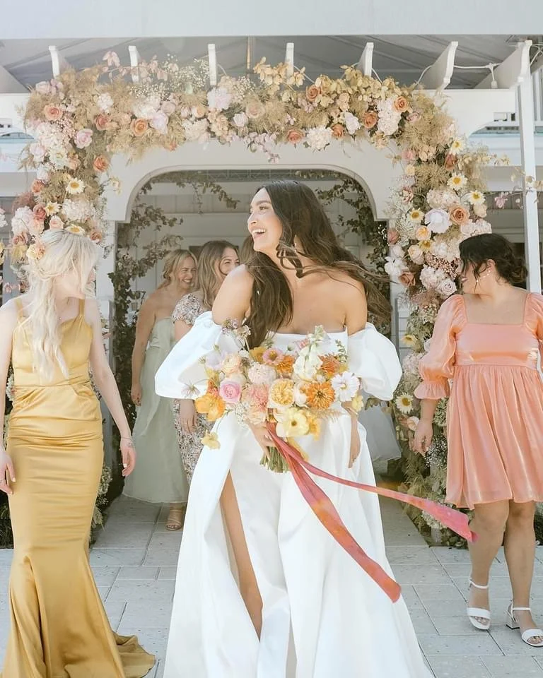 Bride in white dress holding a colorful bouquet, surrounded by bridesmaids in various dresses, walking under a floral arch.