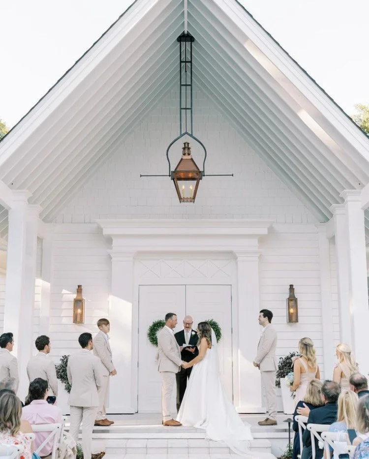 A wedding ceremony taking place outside a white chapel. The couple stands at the altar holding hands, with a priest officiating. Bridesmaids and groomsmen are dressed in light-colored attire, and guests are seated watching the ceremony. The chapel ha