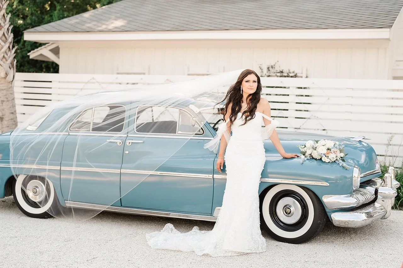 Bride in white dress next to a vintage blue car with a flower arrangement on the hood.