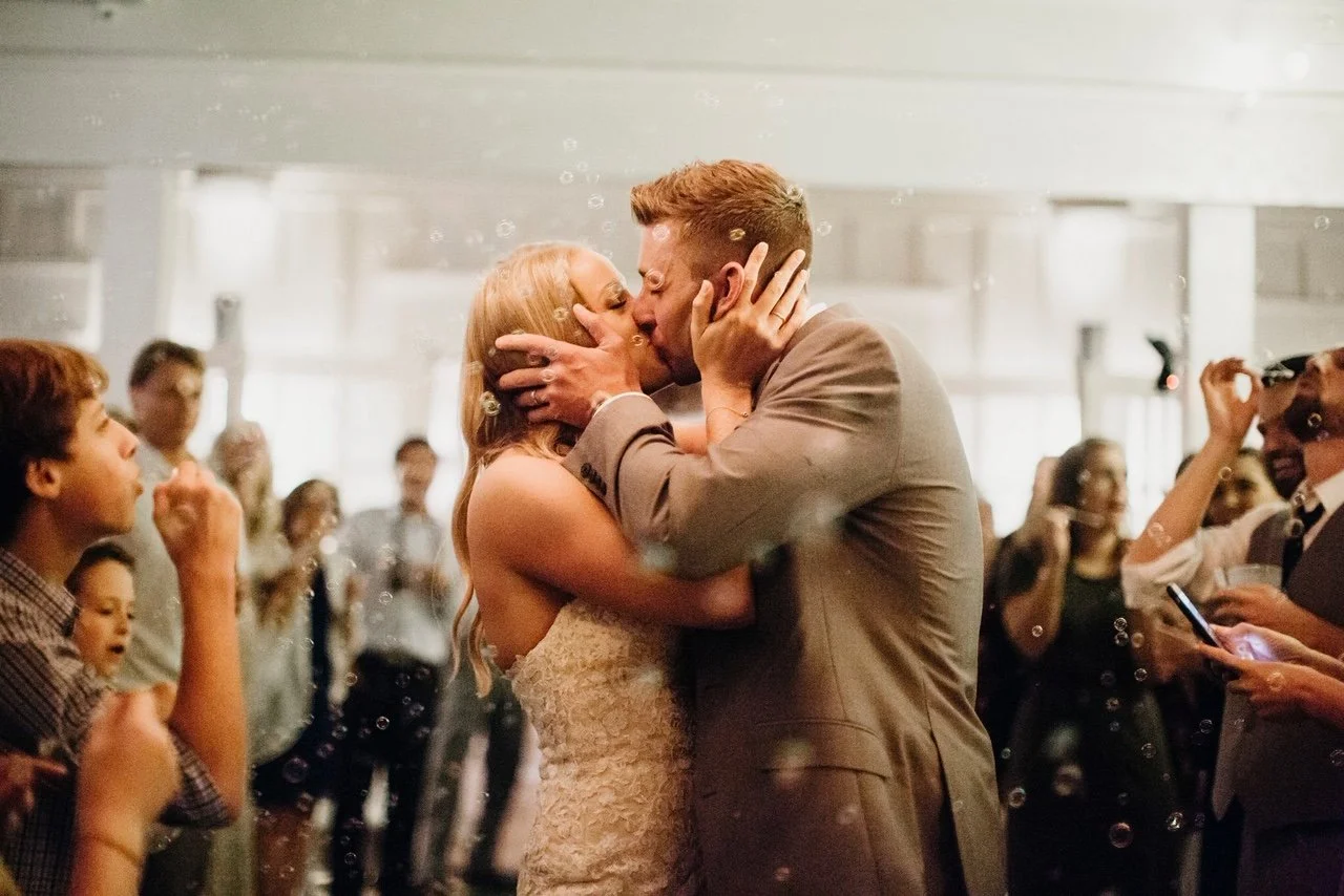 Bride and groom kissing at their wedding with guests in the background, some blowing bubbles.
