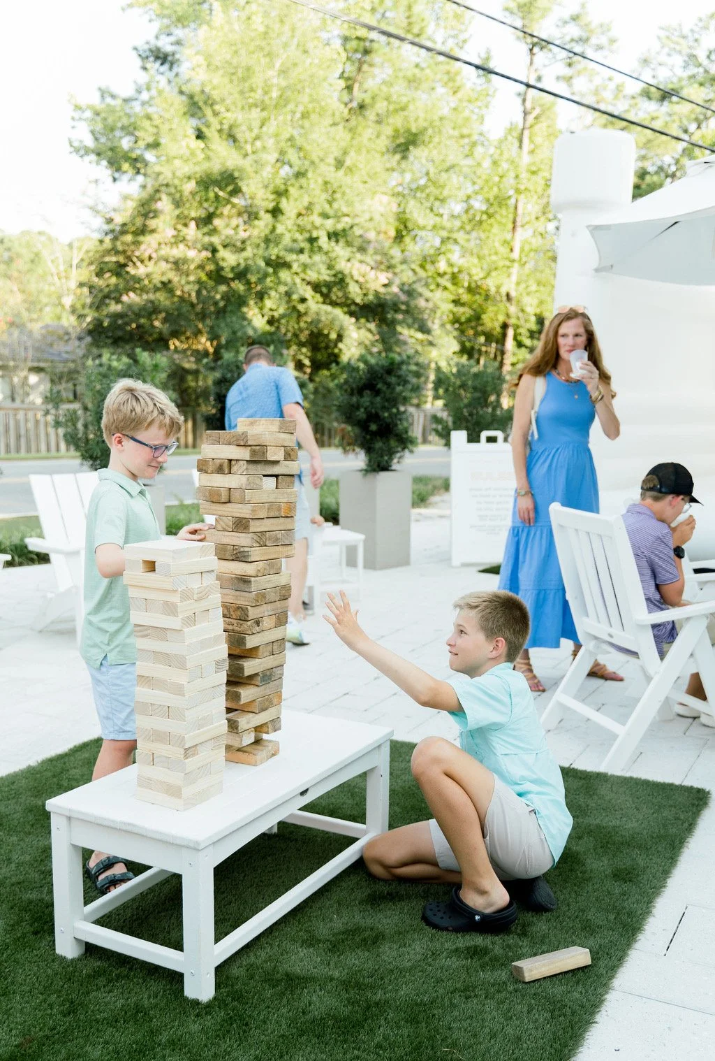 Children playing giant Jenga outdoors, surrounded by adults and greenery.
