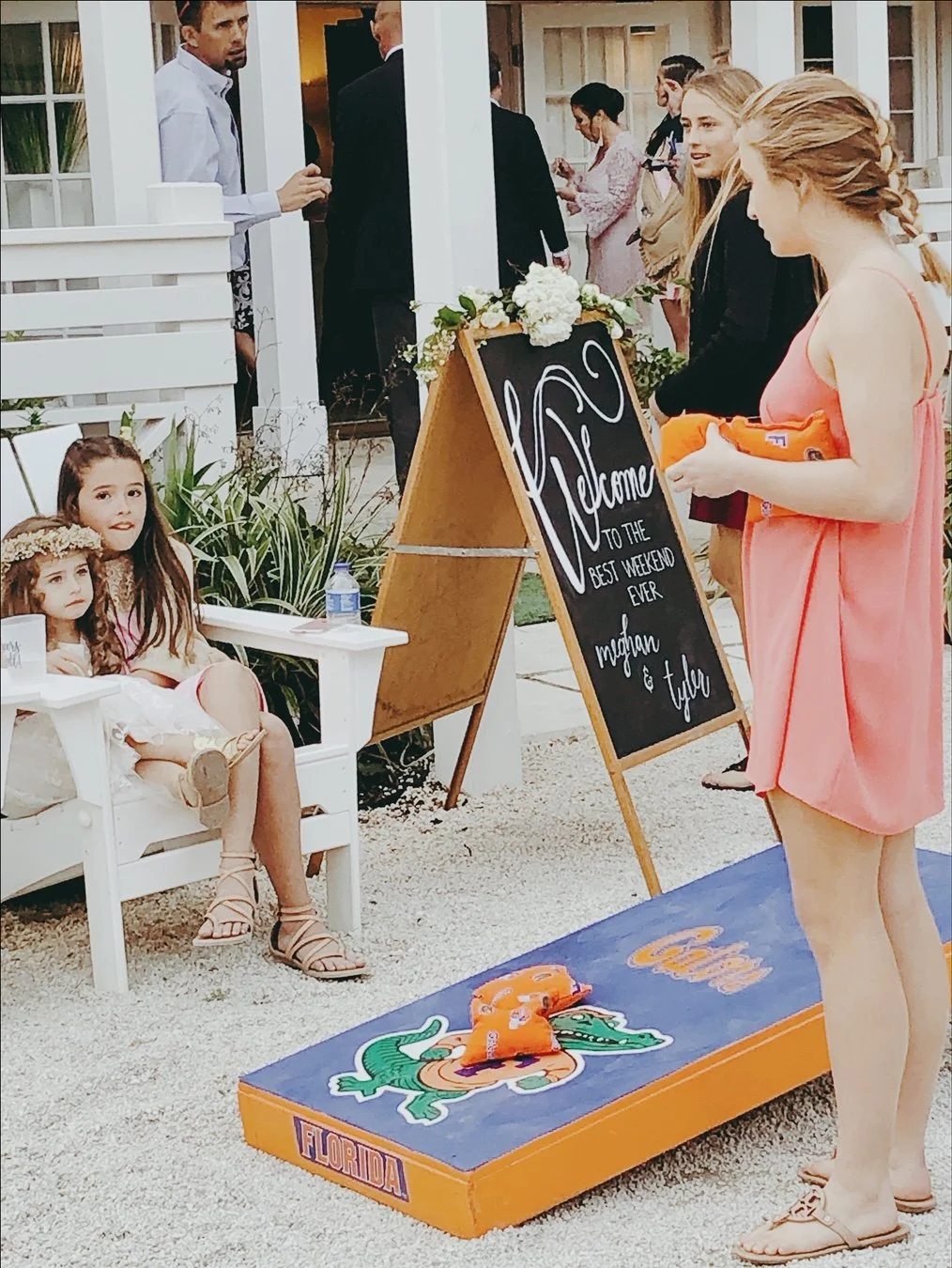 Outdoor event with people and decor featuring a welcome sign, a cornhole game with Florida Gators theme, and seated children.