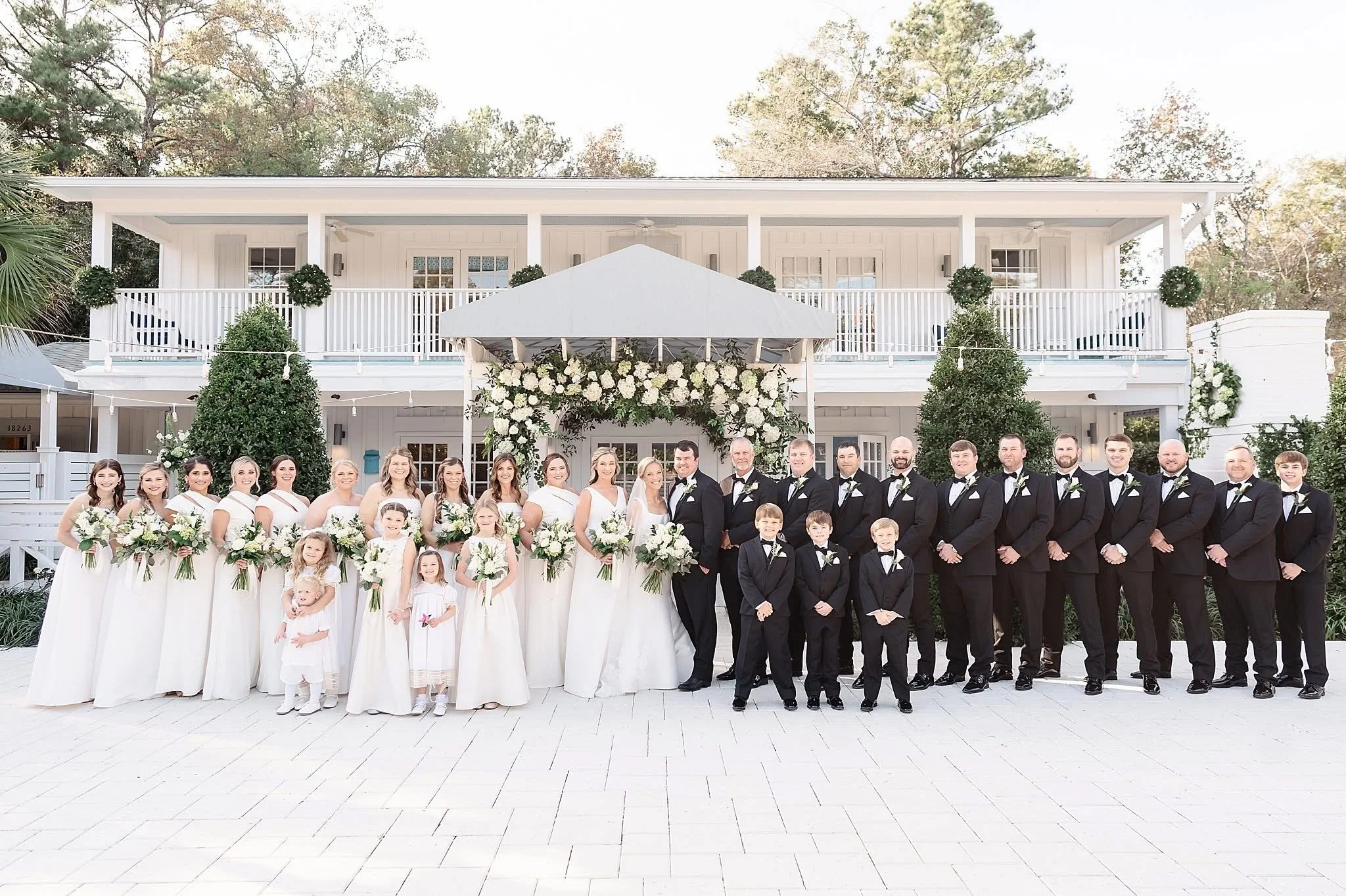 Wedding party group photo in formal attire, with bridesmaids in white dresses and groomsmen in black tuxedos, taken in front of a large white house with floral decorations and greenery.