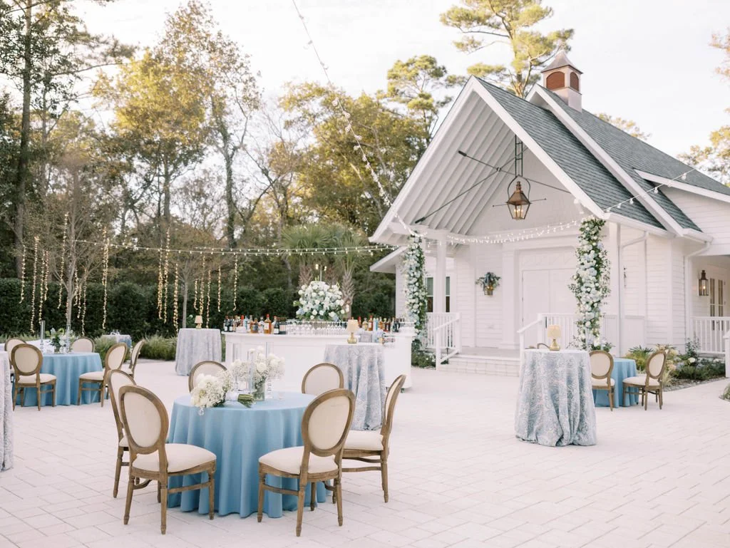 Outdoor wedding reception setup with round tables, blue tablecloths, wooden chairs, and a decorated bar in front of a white chapel. Twinkle lights and floral arrangements adorn the area.