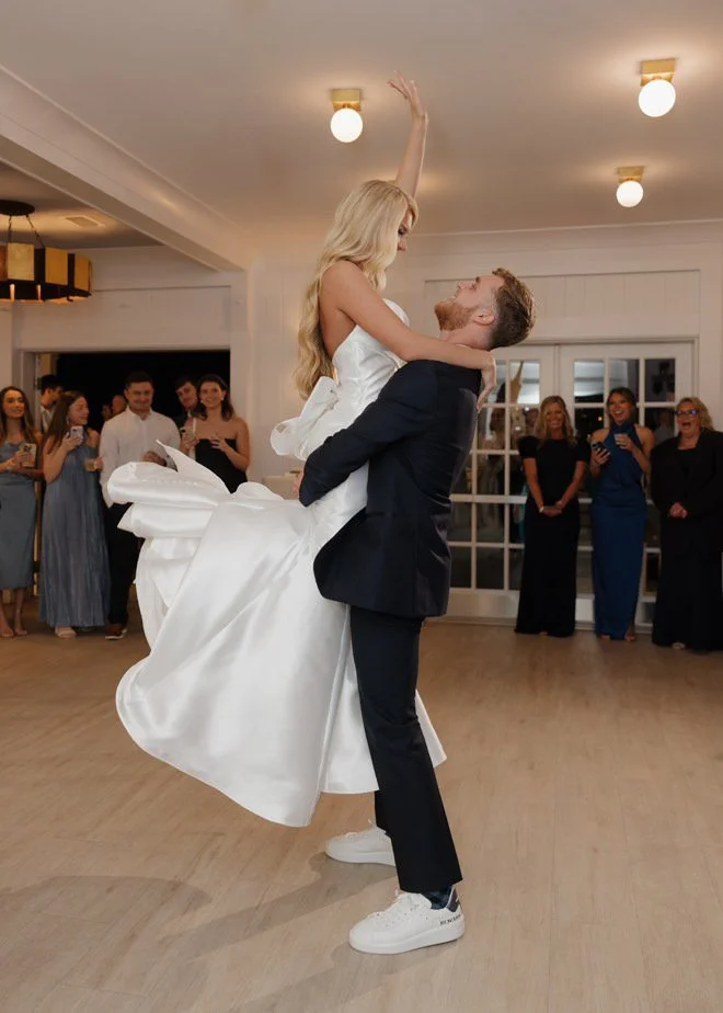 Bride in white dress lifted by groom in dark suit during wedding dance, surrounded by guests indoors.