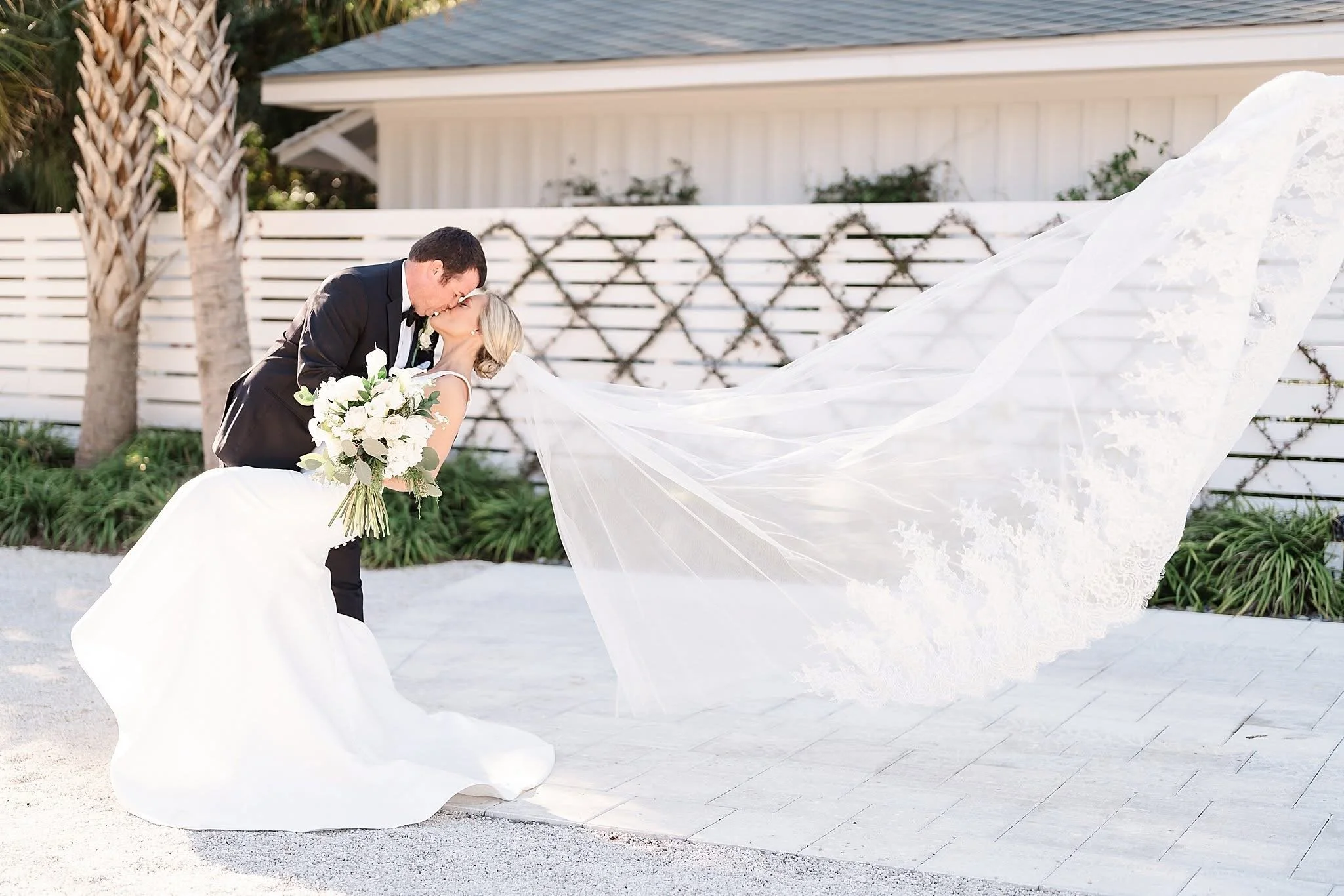 Bride and groom kissing outdoors, bride holding bouquet and wearing flowing veil, groom in black suit, white fence and palm tree in background.